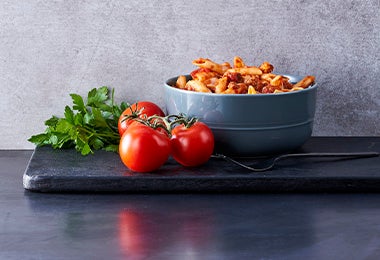 Pasta a la boloñesa junto a tres tomates maduros en un fondo grisáceo y negro.